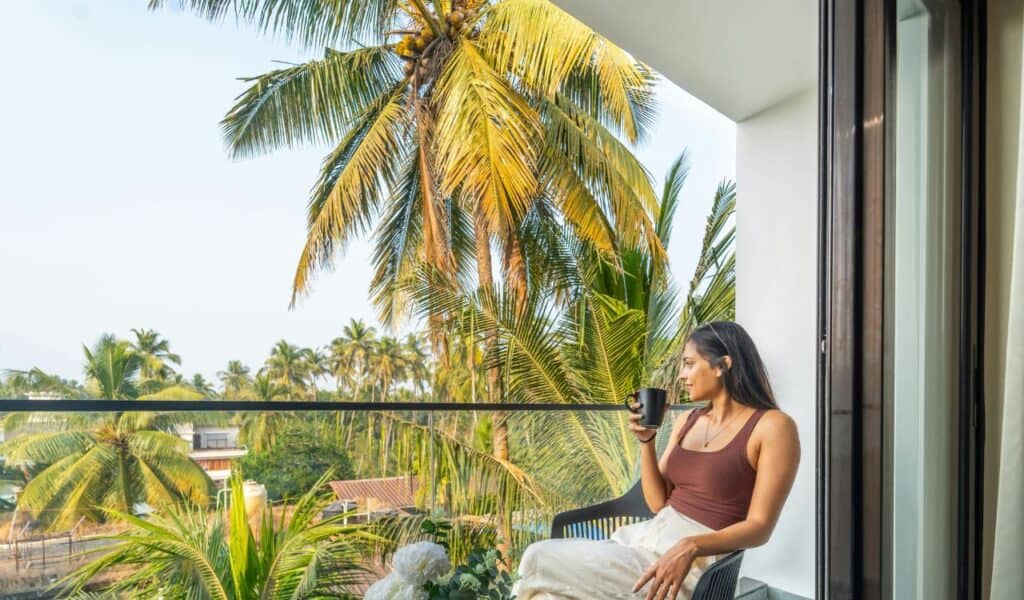 Woman sitting on a balcony with a cup of coffee, surrounded by tropical palm trees and overlooking rooftops in a sunny setting.