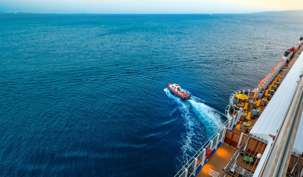 A small red pilot boat sails alongside a large cruise ship in open blue waters, with the ship's deck showing yellow parasols and seating areas.