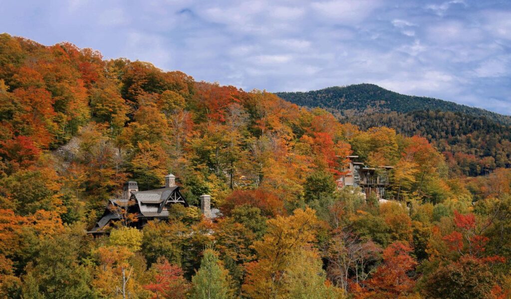 A scenic hillside in Stowe, Vermont, displays vibrant autumn foliage in shades of red, orange, and gold, with charming mountain lodges nestled among the trees and forested peaks in the background.
