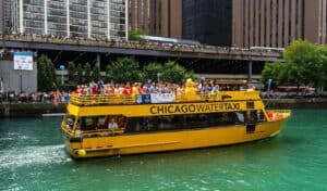A bright yellow Chicago Water Taxi filled with passengers travels along the Chicago River, with crowds gathered on the riverbank and bridge in the background.