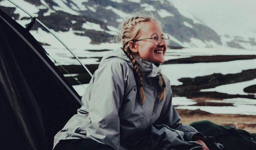 A smiling woman with braided hair and glasses sits outside a tent, dressed in a gray jacket, with a snowy mountain landscape in the background.