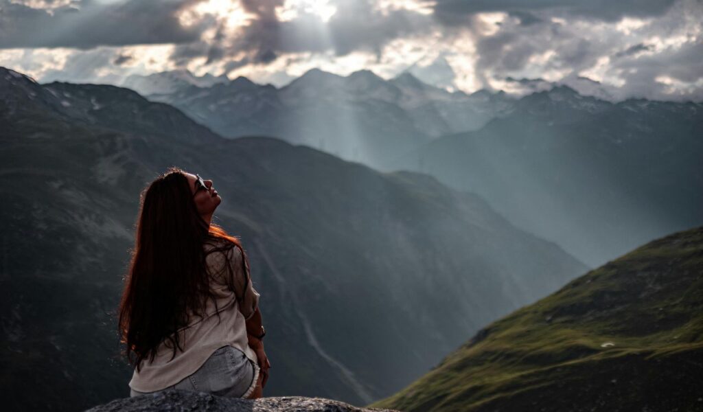 A woman sits on a rock in the Swiss Alps, gazing toward dramatic mountain peaks as rays of sunlight break through the clouds.