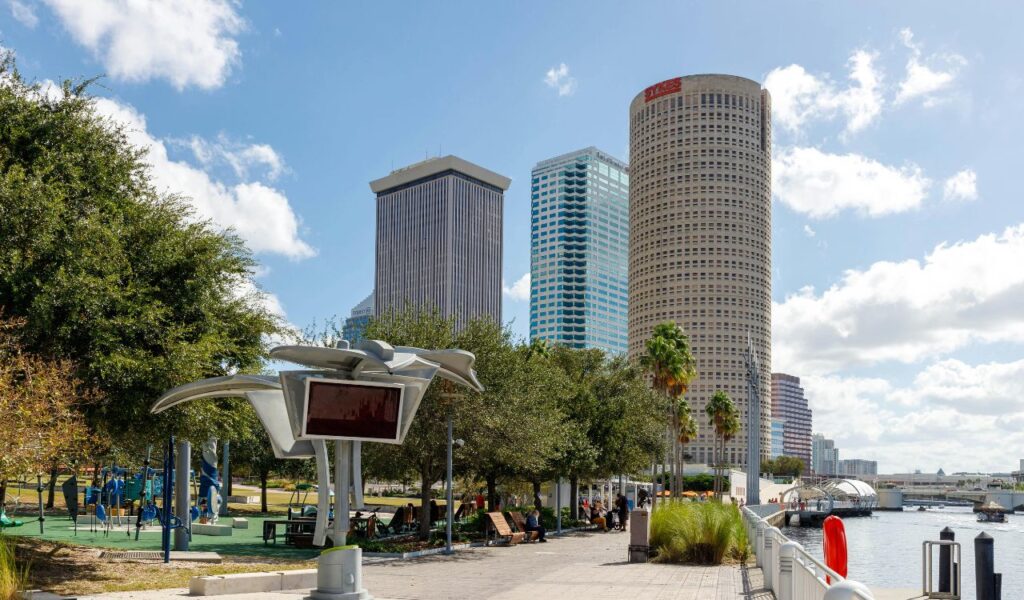 A sunny view of the Tampa Riverwalk with modern high-rise buildings in the background, a tree-lined pedestrian path, public seating areas, and a waterfront with boats docked along the river.