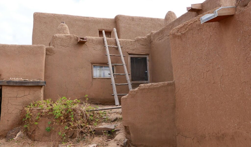 Adobe-style building in Taos, New Mexico, with thick earthen walls, a wooden ladder leading to the upper level, and rustic wooden doors and windows, reflecting traditional Pueblo architecture.