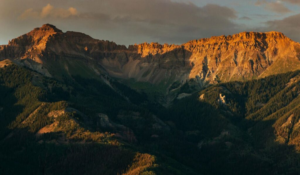 Golden light hitting rugged mountain peaks near Telluride, Colorado, with forested slopes and dramatic cliffs under a partly cloudy sky.