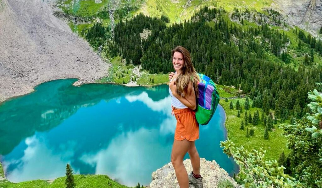 Smiling hiker standing on a rocky ledge overlooking a turquoise alpine lake surrounded by lush green forest and mountains near Telluride, Colorado.