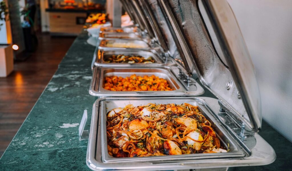 A buffet setup with multiple stainless steel trays of hot food kept warm under lids, showcasing proper temperature control for safe serving.