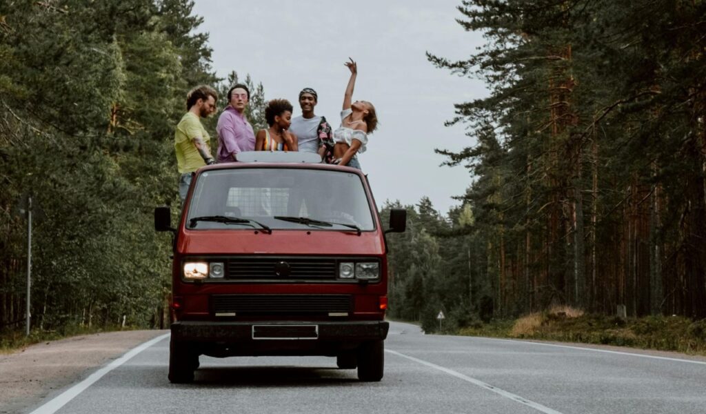 A group of friends enjoying a road trip stands through the sunroof of a red vintage van driving along a forested highway.