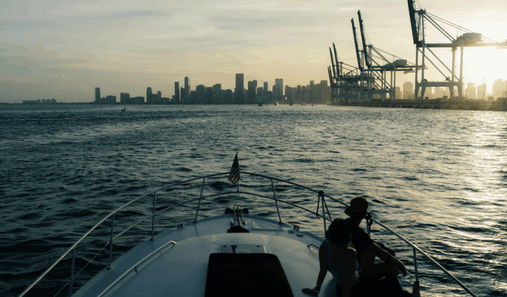 View from the bow of a boat approaching a port city at sunset, with two passengers sitting at the front and large shipping cranes visible on the right against the city skyline.