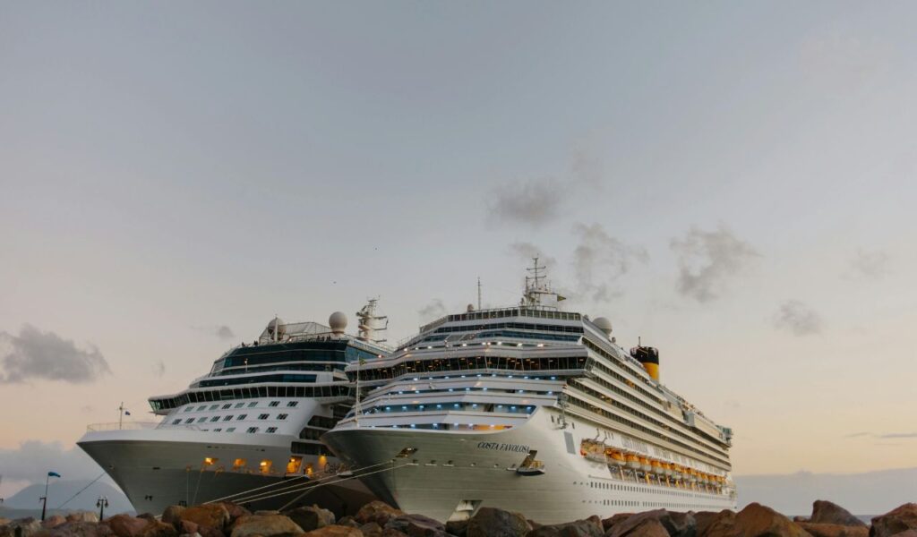 Two large cruise ships docked side by side at a port during early evening, with their lights glowing against a cloudy sky and calm sea.