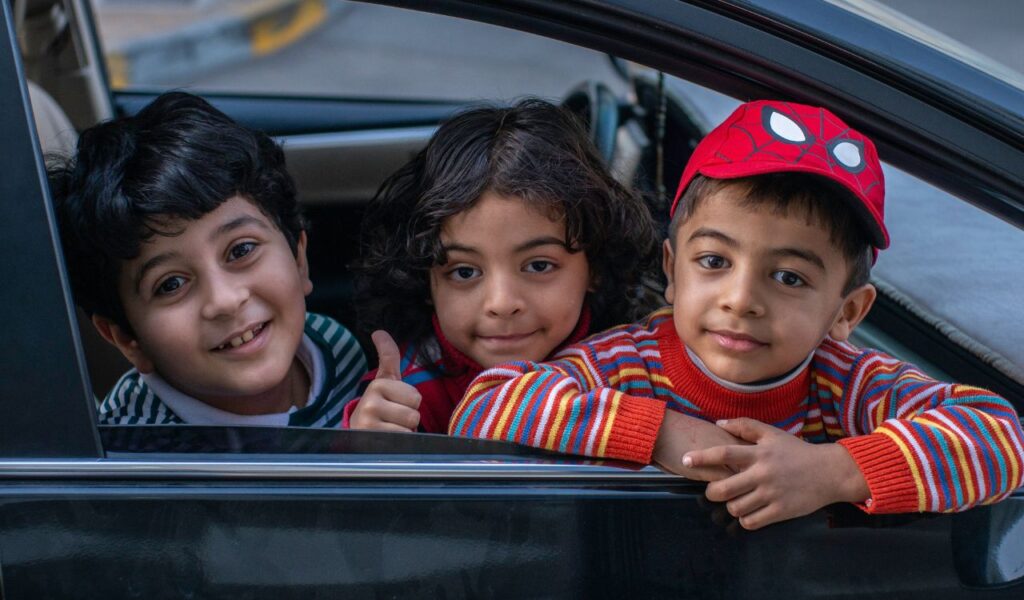 Three smiling kids lean out of a car window, with one giving a thumbs-up and another wearing a red Spiderman cap.