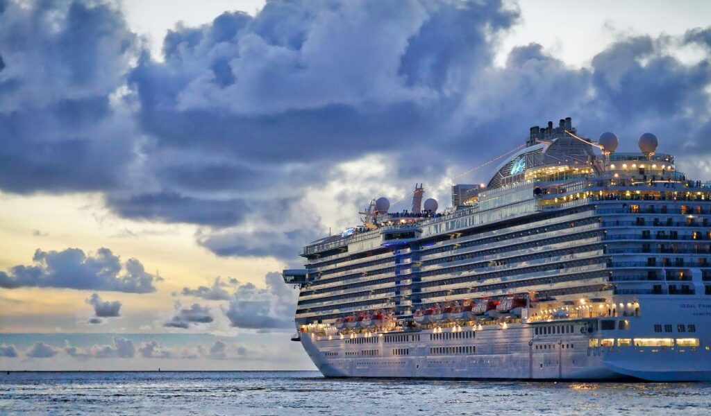 A large cruise ship sails across the ocean at sunset under dramatic clouds, its decks brightly lit while passengers enjoy the view.
