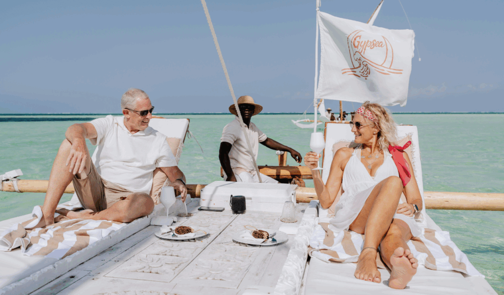 A smiling couple relaxes on lounge chairs aboard a wooden boat in turquoise waters, enjoying drinks and a meal, while a crew member stands at the helm under a sail marked “Gypsea.” A smiling couple relaxes on lounge chairs aboard a wooden boat in turquoise waters, enjoying drinks and a meal, while a crew member stands at the helm under a sail marked “Gypsea.” A smiling couple relaxes on lounge chairs aboard a wooden boat in turquoise waters, enjoying drinks and a meal, while a crew member stands at the helm under a sail marked “Gypsea.