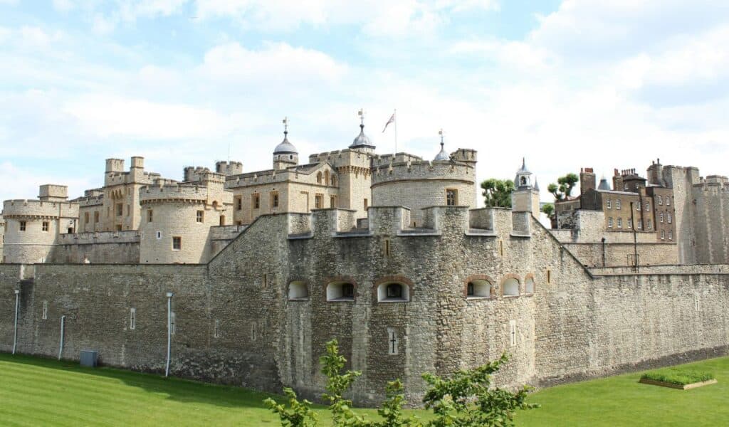 The Tower of London in England, a historic fortress with stone walls, turrets, and battlements, set against a partly cloudy sky with a well-kept green lawn in the foreground.
