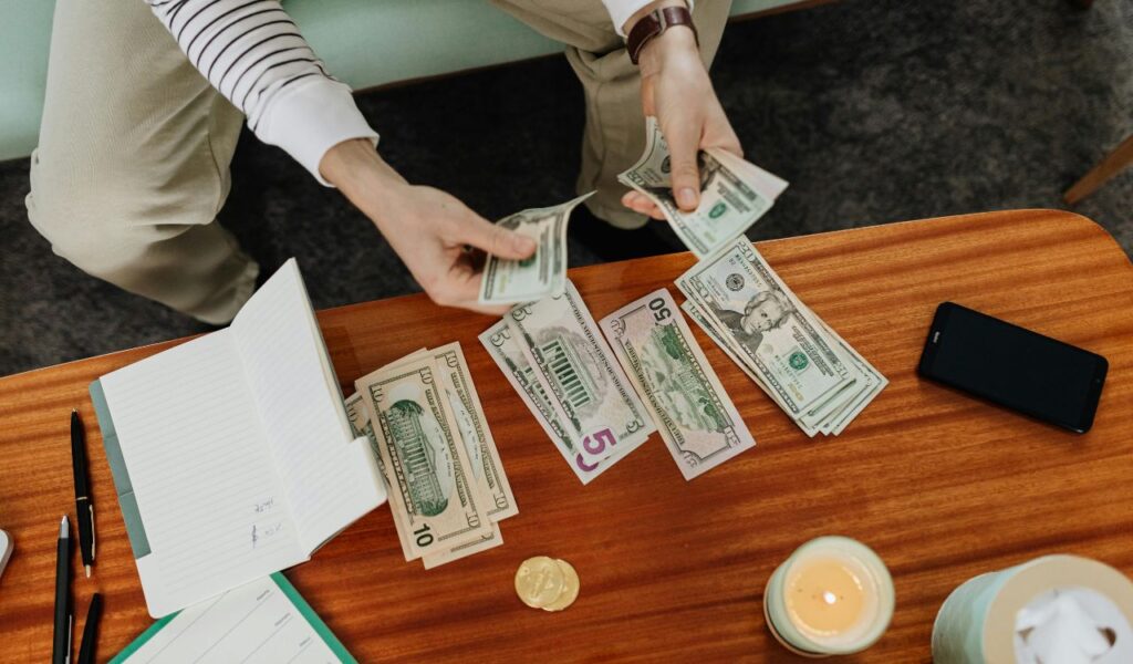 Person counting US dollar bills at a wooden table with a notebook, pens, a candle, and a smartphone, representing budgeting or tracking expenses.