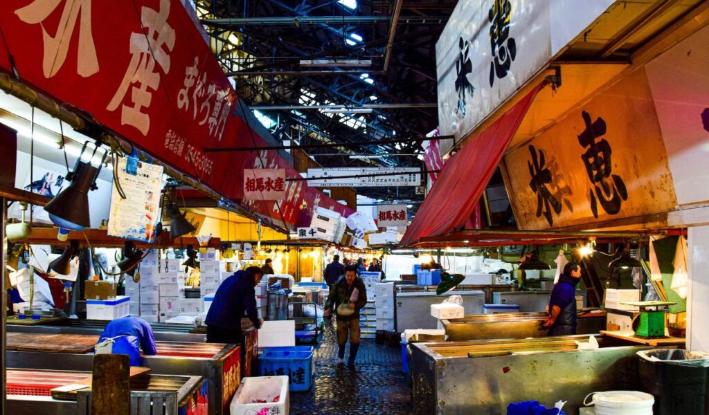 Vendors working at stalls in Tsukiji Outer Market in Tokyo, with red and white signs displaying Japanese characters overhead.