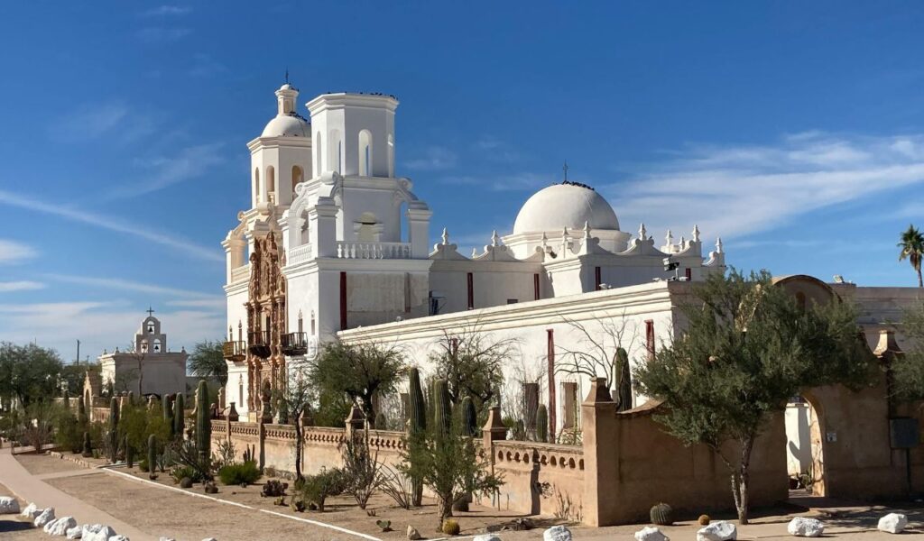 Historic Mission San Xavier del Bac in Tucson, Arizona, featuring white Spanish colonial architecture, domes, and towers surrounded by desert landscaping and cacti under a clear blue sky