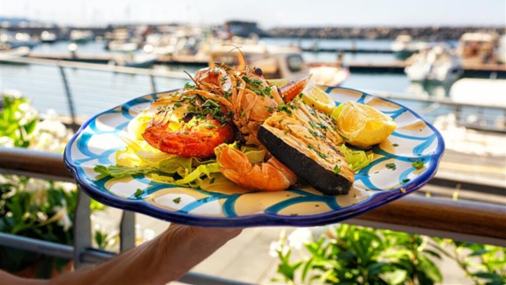 Serving Seafood and fish lunch in sea coast restaurant, Sicily, Italy. Italian food. Outdoor. Port with yacht on background