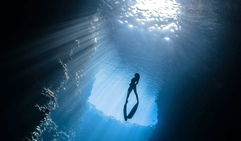 A lone diver glides upward in a deep underwater cave, illuminated by dramatic rays of sunlight streaming through the water’s surface above.