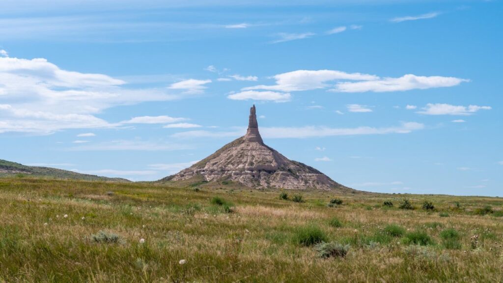 Chimney Rock stands tall against the blue sky in Nebraska during a sunny summer day, surrounded by grassy plains and scattered clouds