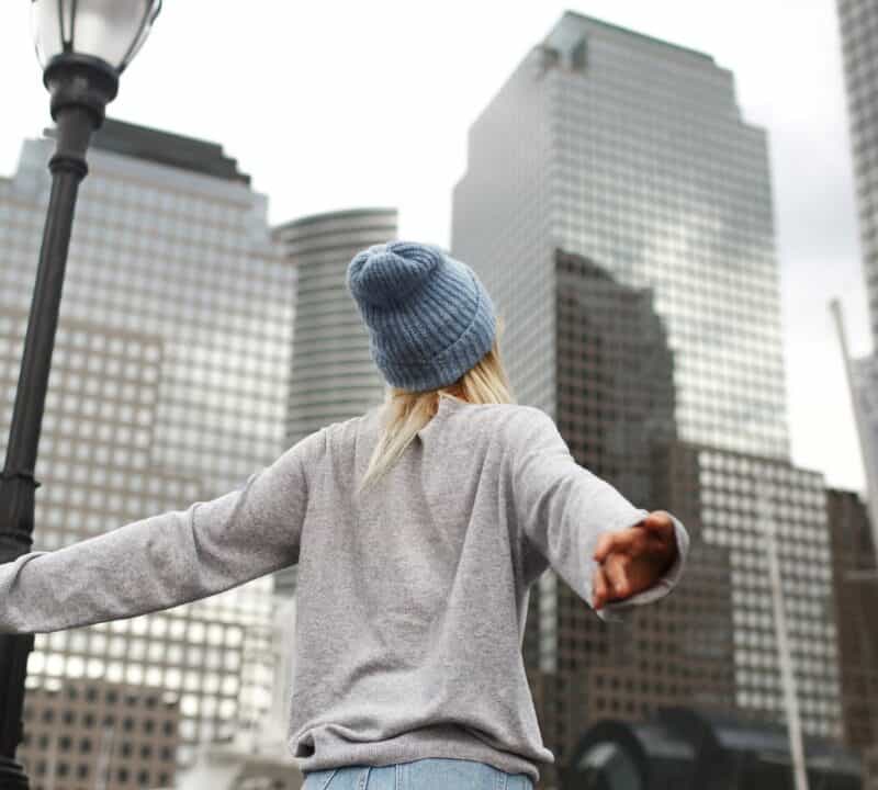 Girl in blue hat and grey sweater stands on the street with skyscrapers