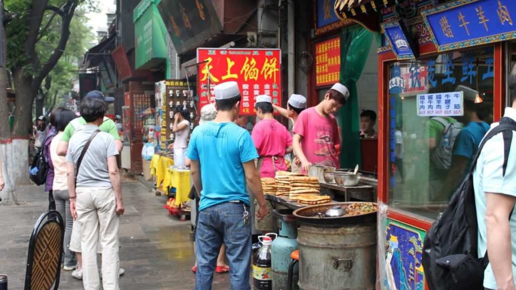 Xi’an, China – June 01, 2011: A street food vendor prepares stacks of freshly cooked flatbreads next to a large pot of sizzling, seasoned meat in a lively market setting