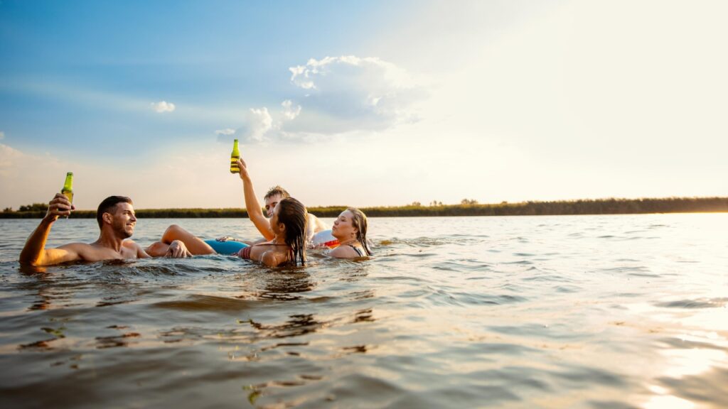 Group of young friends having fun and enjoying a summer day swimming with rubber tube and drinking beer at the lake.