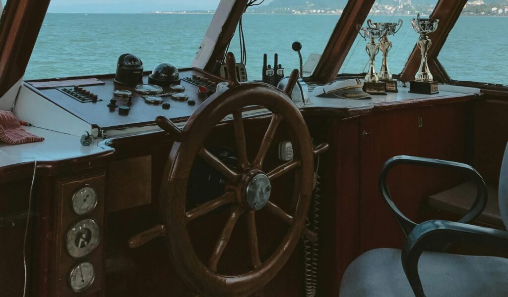 Wooden ship steering wheel inside a boat's control room with gauges, radios, and trophies overlooking the sea