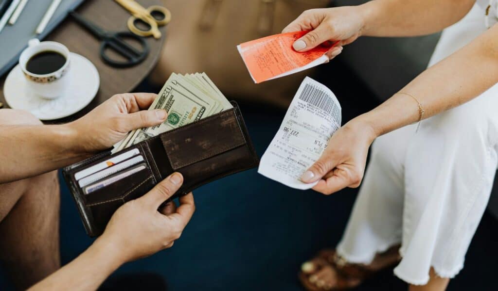 Person holding a wallet with cash and cards while another person hands over receipts and a small ticket.