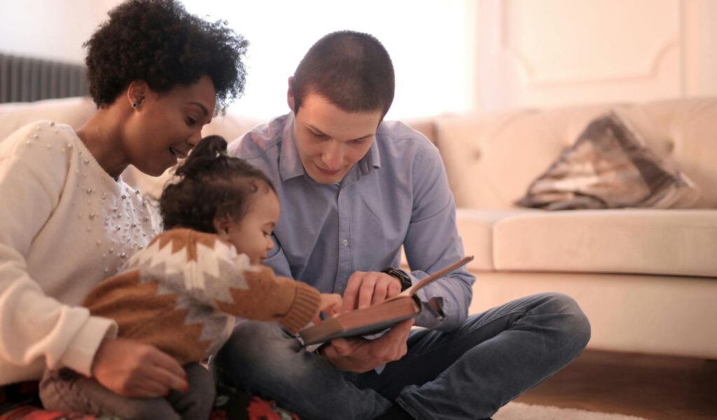Smiling parents sitting on the floor with their young child, using a book as a prop to engage and interact together.