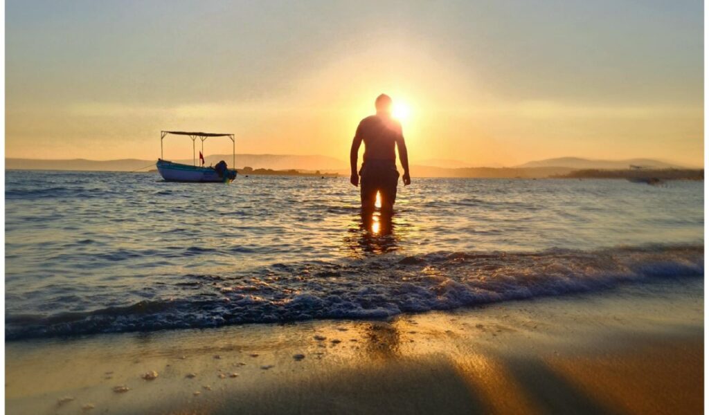 Silhouette of a person standing in shallow water at sunrise, with a small boat floating nearby and mountains in the distance.