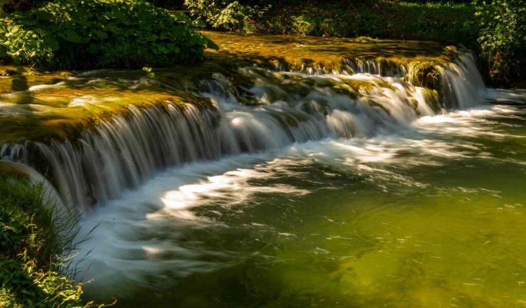 A serene waterfall flowing gently over moss-covered rocks into a clear green pool at Wakulla Springs, Florida, surrounded by lush greenery