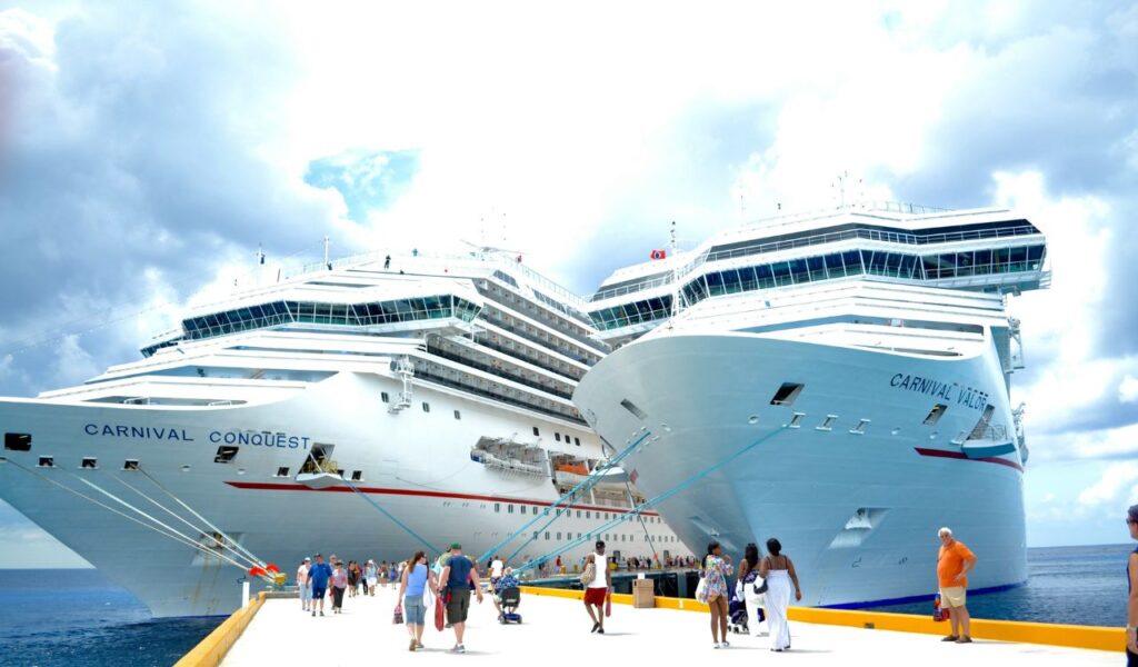 Two large Carnival cruise ships, Carnival Conquest and Carnival Valor, docked side by side at a pier with passengers walking along the dock under a partly cloudy sky.


