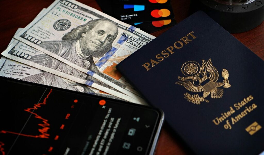Close-up of U.S. $100 bills, an American passport, business debit cards, and a smartphone displaying a stock market graph