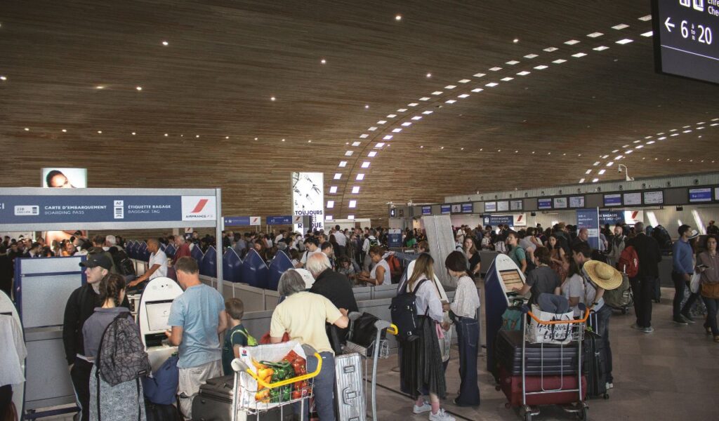 Crowded airport check-in area with travelers waiting in long lines, some with luggage carts and suitcases.