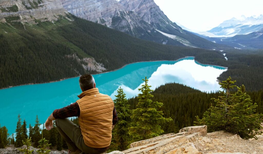A man in an orange vest sits on a rocky ledge overlooking a turquoise lake surrounded by dense evergreen forests and towering mountains.