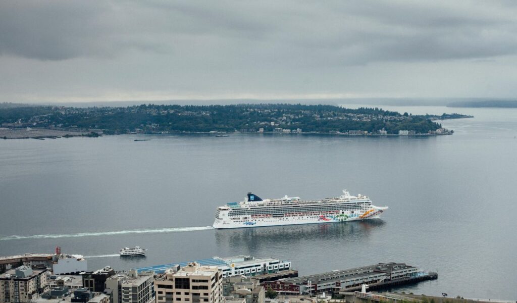 A large Norwegian Cruise Line ship departs from a port, leaving a white wake in the calm gray water, with a small ferry nearby and a coastal city in the background under an overcast sky. A large Norwegian Cruise Line ship departs from a port, leaving a white wake in the calm gray water, with a small ferry nearby and a coastal city in the background under an overcast sky. A large Norwegian Cruise Line ship departs from a port, leaving a white wake in the calm gray water, with a small ferry nearby and a coastal city in the background under an overcast sky.