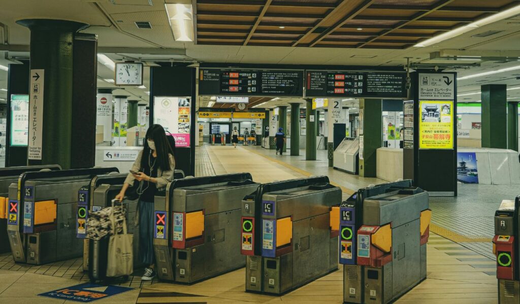 Woman wearing a mask pulls luggage through a ticket gate at a train station while looking at her phone.
