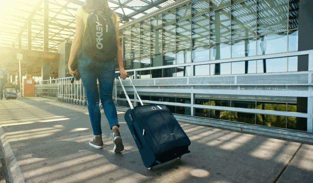 A traveler walking outside an airport terminal, pulling a rolling suitcase and carrying a black Oakley backpack, with sunlight streaming through the glass and metal roof structure.