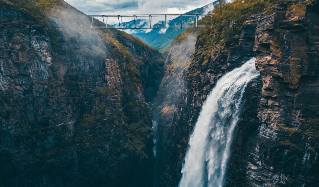 A dramatic waterfall cascades down a steep rocky cliff into a deep gorge, while a narrow pedestrian bridge spans high above the canyon, connecting the cliffs with mist rising into the mountainous backdrop.
