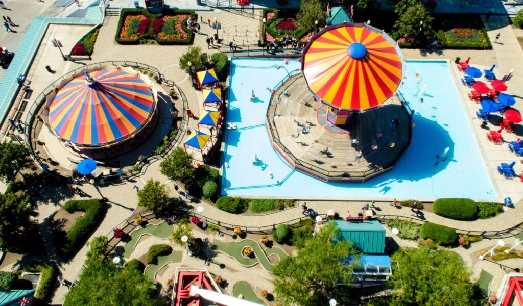 A vibrant aerial view of the Wisconsin State Fair shows colorful striped tents, a carousel over water, mini-golf, and shaded seating areas.