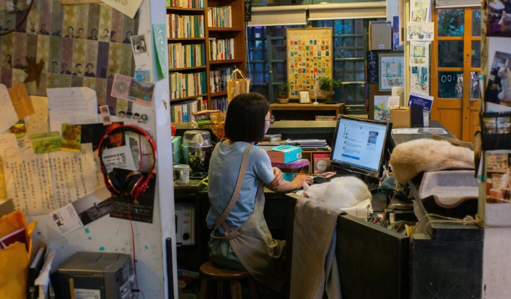 A cozy bookstore interior with a woman seated at a desk using a computer, surrounded by bookshelves, notes, and a cat resting nearby.