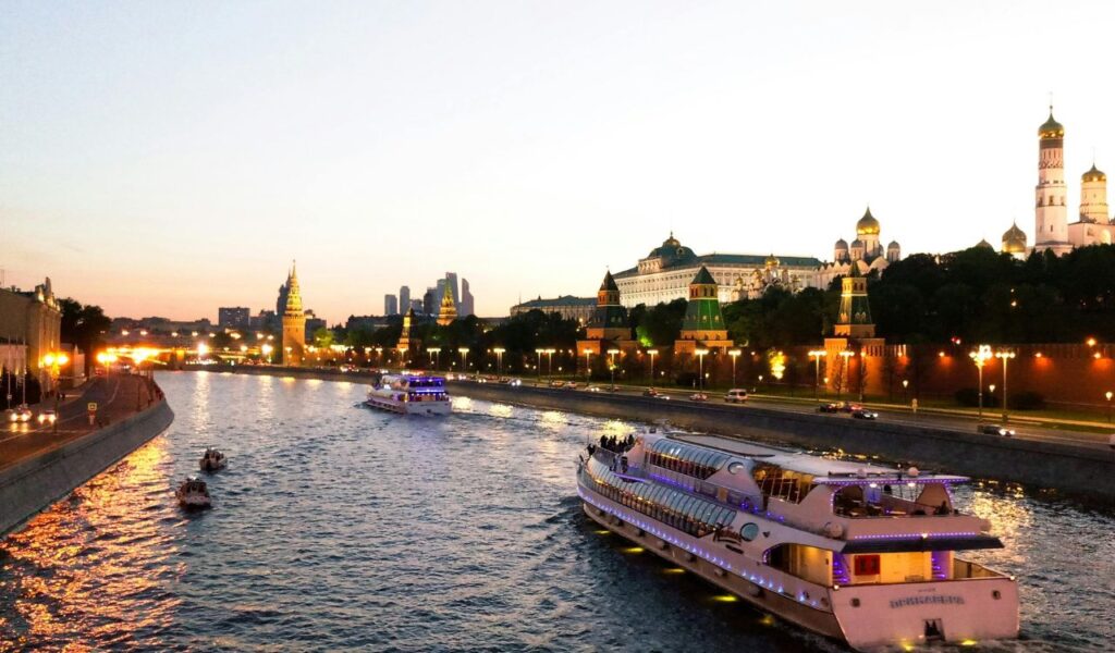 Illuminated river cruise boats sailing through a city at dusk, with historic buildings and domed towers glowing under a twilight sky