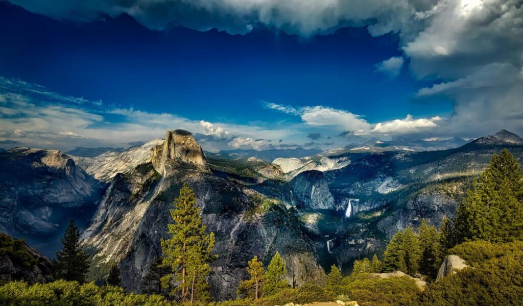 A stunning view of Yosemite National Park featuring Half Dome, surrounding granite cliffs, dense pine forests, and a distant waterfall under a dramatic cloudy sky.