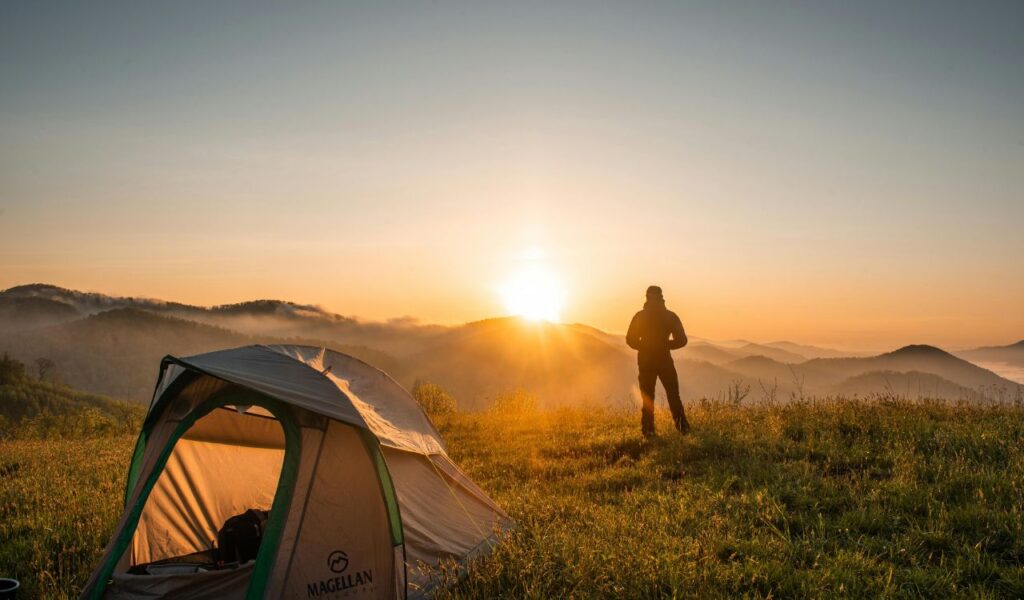 Camper stands beside a tent on a grassy hilltop at sunrise, overlooking misty mountain ranges and glowing skies in Yosemite Valley, California.