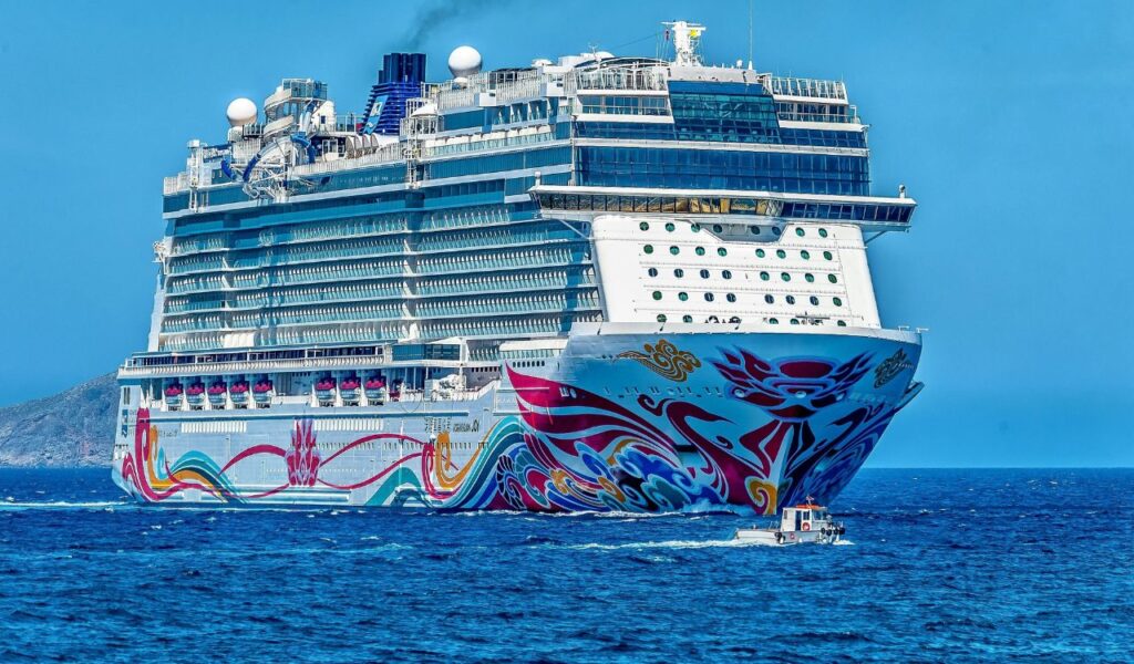 A large, colorful cruise ship sailing on the ocean with a small boat nearby and land visible in the background.