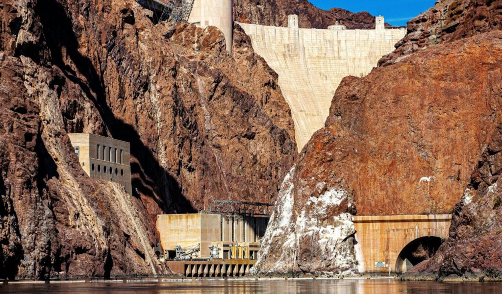View of Hoover Dam surrounded by rugged cliffs with power station buildings and a tunnel entrance at the base near the water.