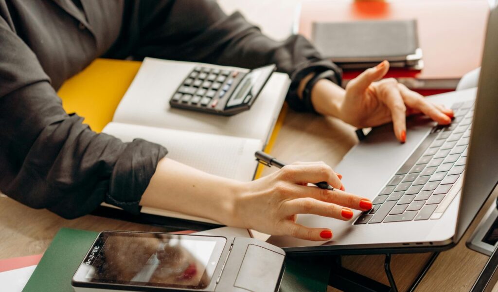 Close-up of a person working on a laptop with a calculator, notebook, and smartphone on the desk, managing finances and expenses.