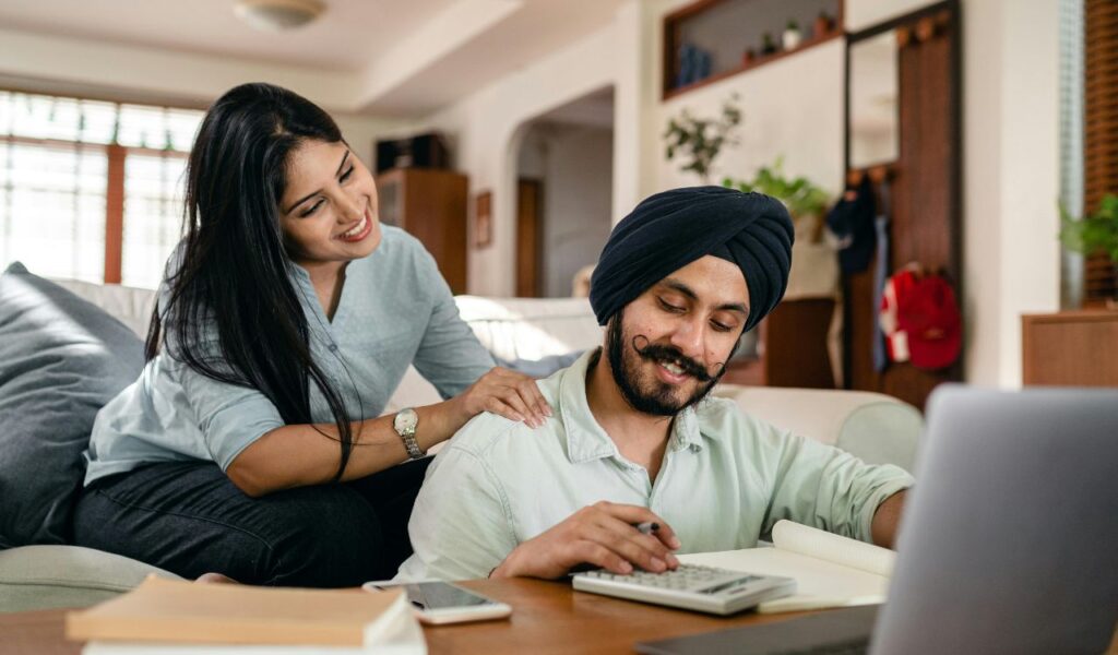 A smiling couple sitting together at home, discussing finances while using a calculator and laptop, planning their budget.