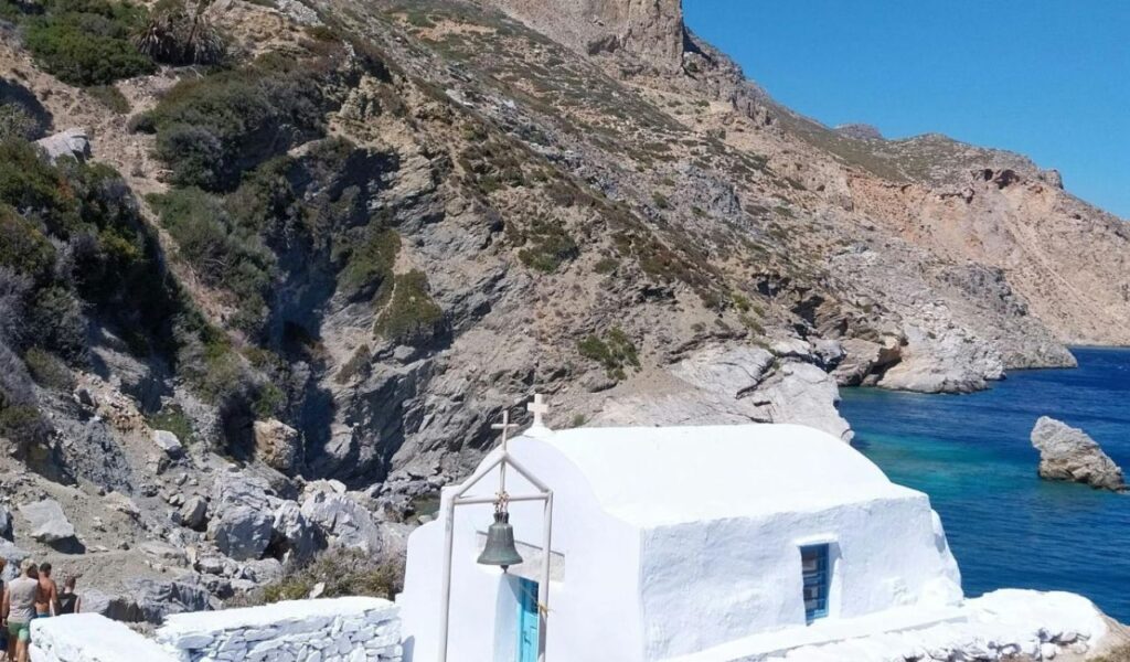 A small whitewashed chapel with a blue door and bell stands by the rocky coastline of Amorgos, Greece, overlooking the deep blue Aegean Sea.